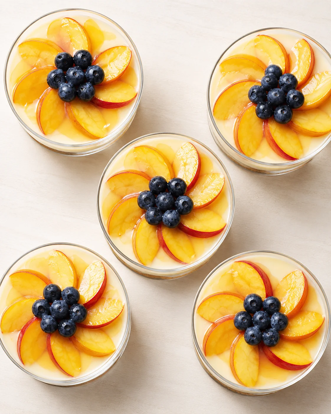 Glass dessert cups filled with peach slices arranged in a flower pattern around blueberries atop creamy yogurt
