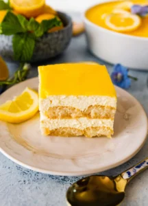 Square slice of lemon-layered cake with bright yellow glaze on a marble plate, lemon wedges nearby.