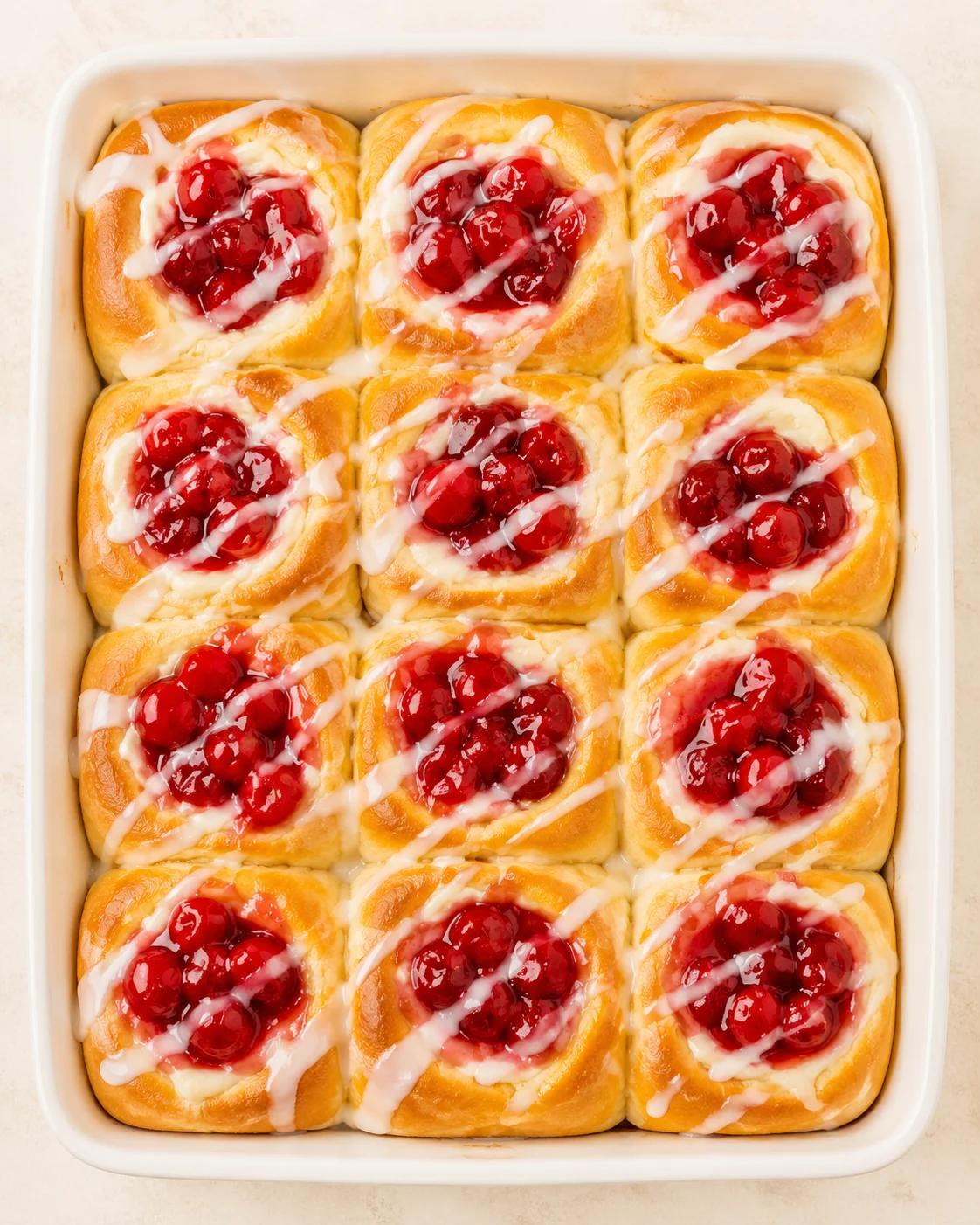 Top-down view of a white baking dish filled with nine cherry-filled cinnamon buns, drizzled with white icing.