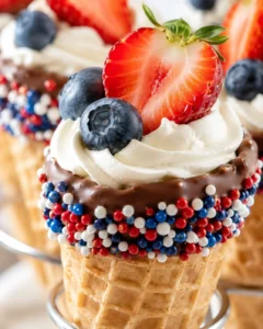 Close-up of a chocolate-dipped cone with whipped cream, strawberry slice, blueberries, and red-white-blue sprinkles.