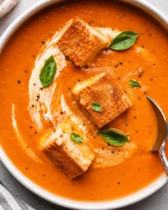 A bowl of creamy, roasted tomato soup garnished with basil leaves and croutons.