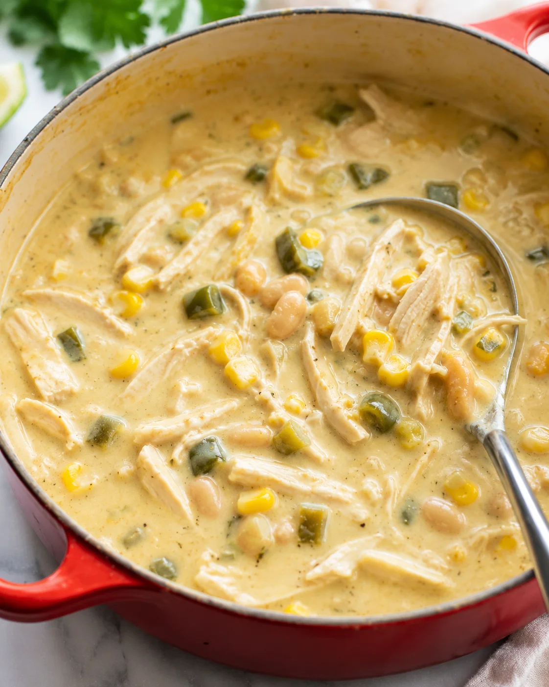 A hearty, creamy white chicken chili with shredded chicken, corn, and green chilies in a red pot.