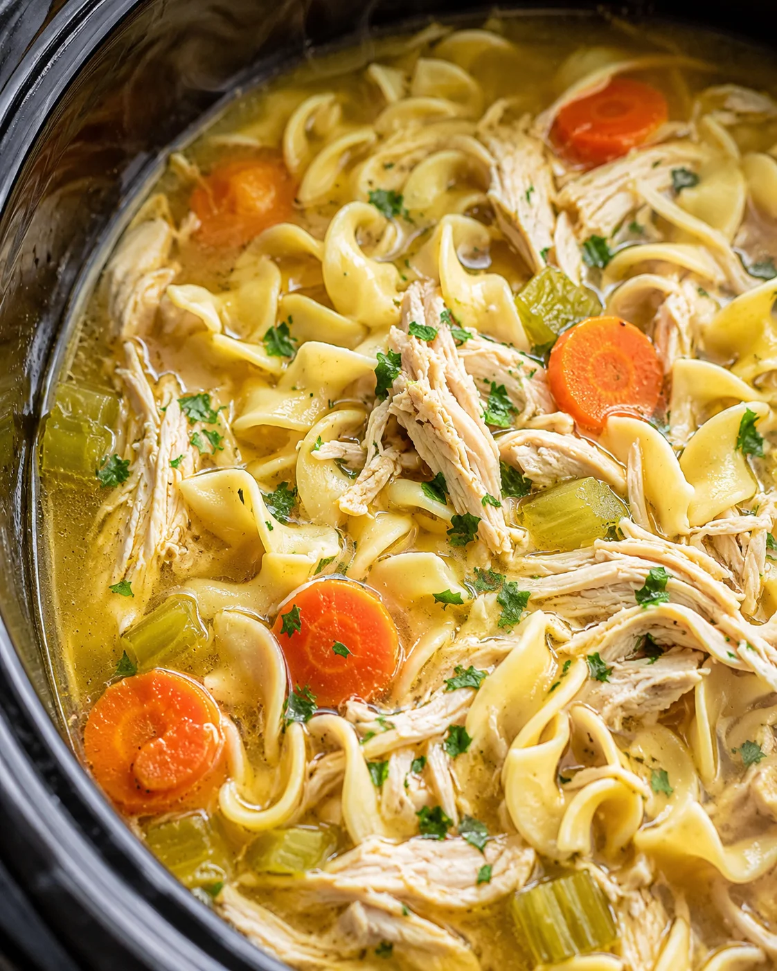 Close-up of homemade chicken noodle soup in a slow cooker with vegetables and shredded chicken.