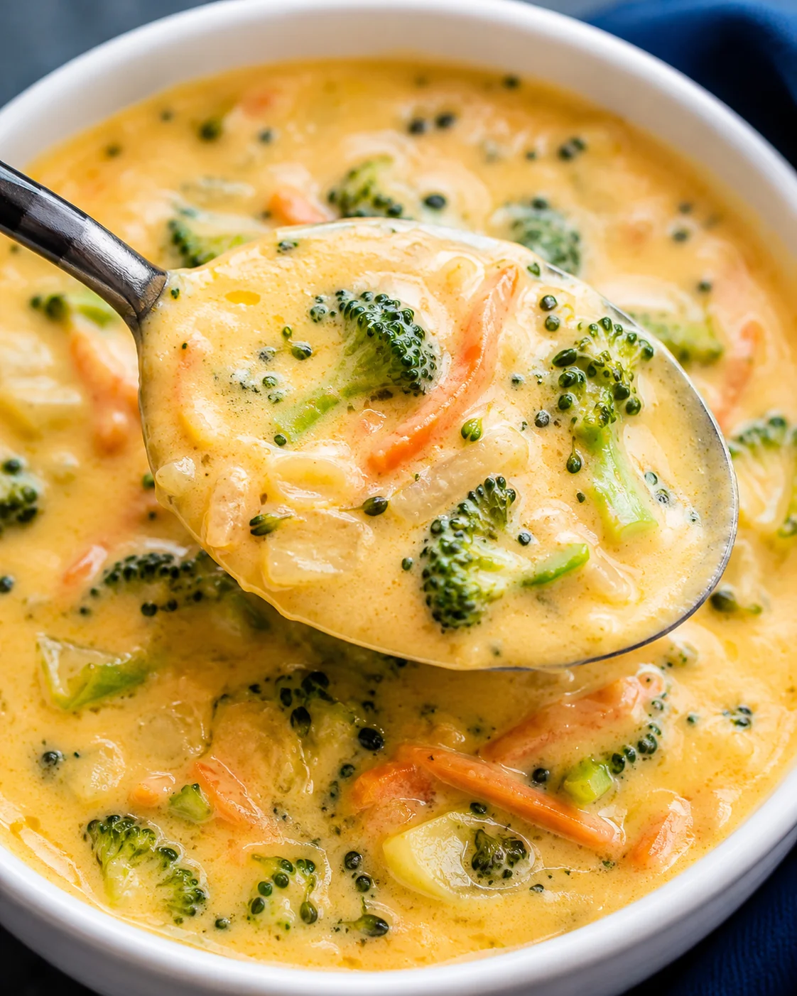 A spoonful of rich broccoli cheddar soup with vegetables and cheese in a white bowl.
