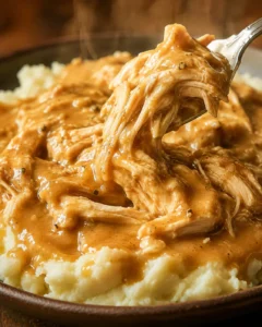 Spoon lifting shredded slow cooker chicken and gravy over mashed potatoes